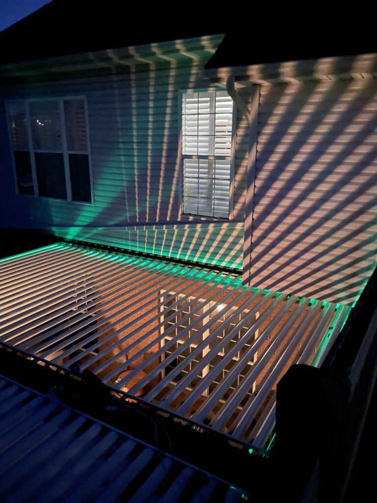 picture of 2 bay bronze sundance louvered pergola with white louvers open and led strip lighting mounted to home